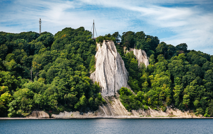 Kreidefelsen an einer Küste mit bewaldetem Hügel und blauem Himmel.