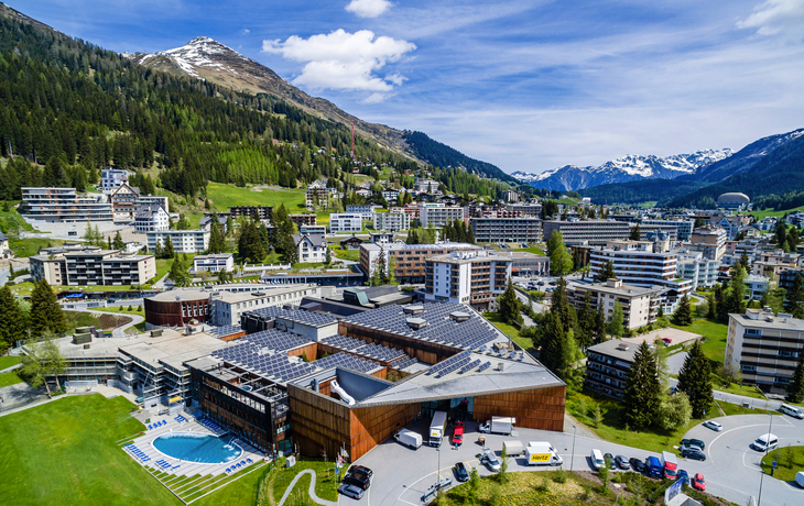 Luftaufnahme von Davos mit Bergen, Gebäuden und bewaldeten Hügeln im Hintergrund.